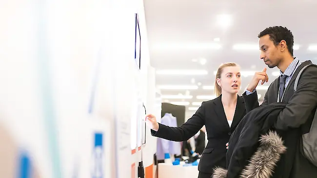 Eine Frau erklärt auf einer Messe einem Mann etwas auf einem großen Bildschirm an einem Messestand. Eine Frau erklärt auf einer Messe einem Mann etwas auf einem großen Bildschirm an einem Messestand.
