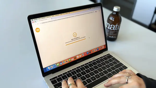 Ein Laptop zeigt auf dem Bildschirm eine Bestätigungsmeldung, während Hände die Tastatur bedienen; im Hintergrund steht eine Getränkeflasche. Ein Laptop zeigt auf dem Bildschirm eine Bestätigungsmeldung, während Hände die Tastatur bedienen; im Hintergrund steht eine Getränkeflasche.