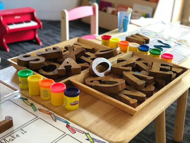 Ein hölzerner Aktivitätstisch in einem Klassenraum mit Schalen großer Holzbuchstaben und bunten Knete‑Töpfen, bereit für kreatives Kinderspiel. Ein hölzerner Aktivitätstisch in einem Klassenraum mit Schalen großer Holzbuchstaben und bunten Knete‑Töpfen, bereit für kreatives Kinderspiel.