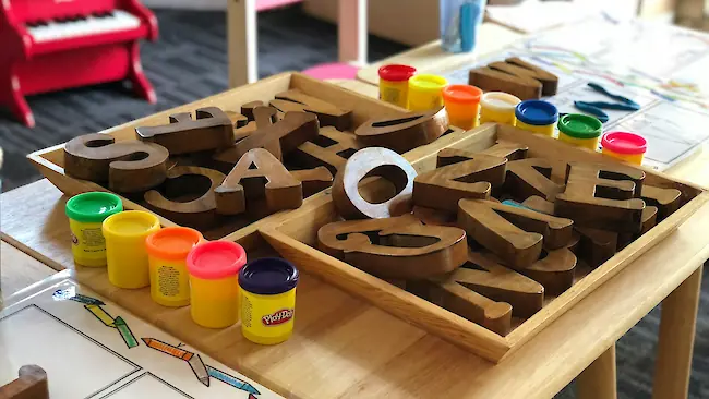 Ein hölzerner Aktivitätstisch in einem Klassenraum mit Schalen großer Holzbuchstaben und bunten Knete‑Töpfen, bereit für kreatives Kinderspiel. Ein hölzerner Aktivitätstisch in einem Klassenraum mit Schalen großer Holzbuchstaben und bunten Knete‑Töpfen, bereit für kreatives Kinderspiel.