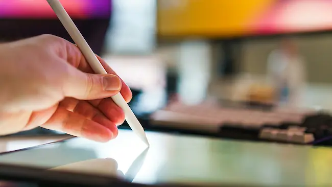 Eine Hand hält einen weißen Stylus und arbeitet am Bildschirm eines Grafiktabletts in einem unscharfen Arbeitsbereich mit Computerbildschirmen im Hintergrund. Eine Hand hält einen weißen Stylus und arbeitet am Bildschirm eines Grafiktabletts in einem unscharfen Arbeitsbereich mit Computerbildschirmen im Hintergrund.